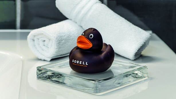 A black rubber duck with an orange beak and the text "Sorell Hotels Switzerland" on a glass table. A white towel is rolled up in the background. -  Sorell Boutique-Hotel City Weissenstein St. Gallen (St. Gallen)