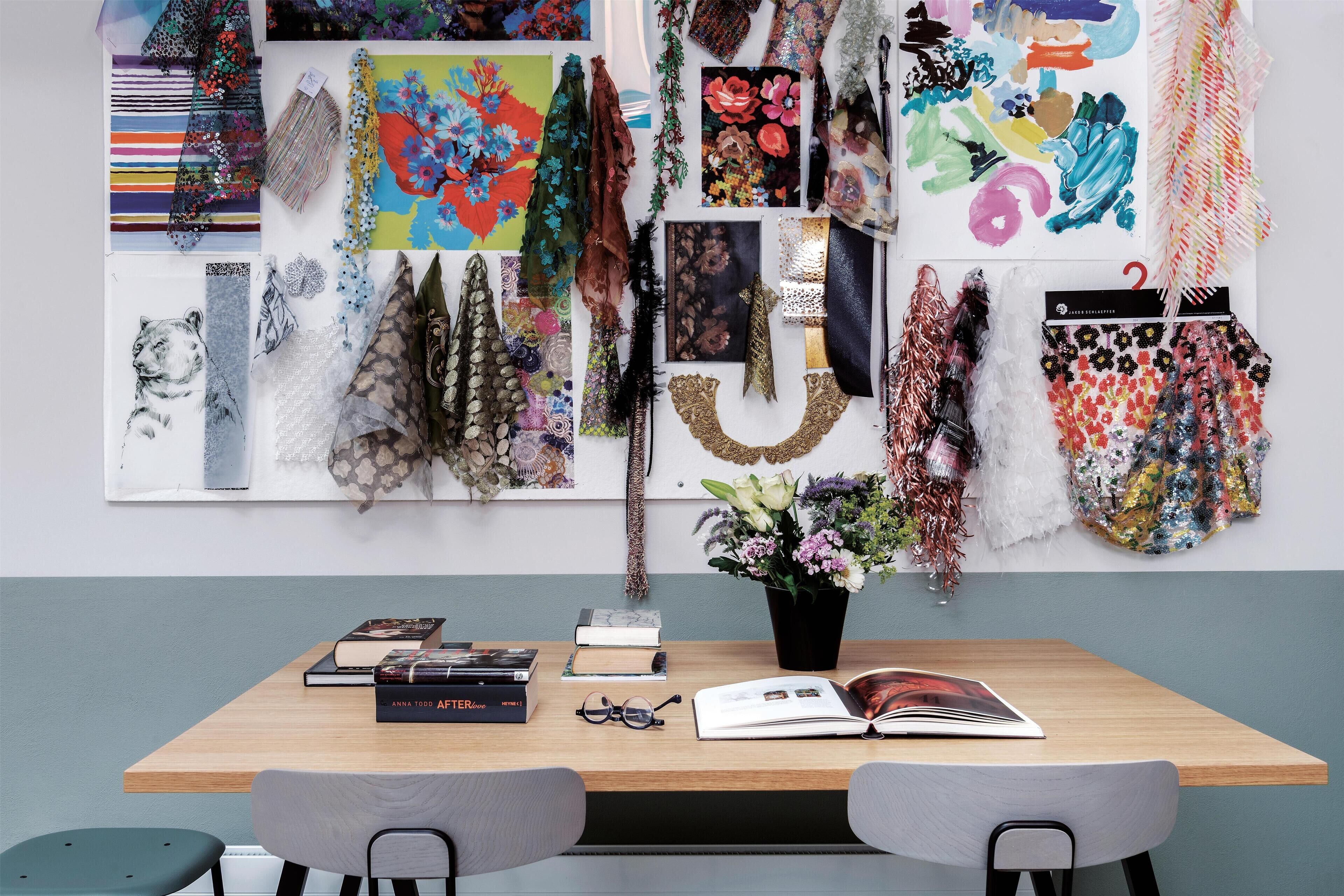 A room with a wooden table, two chairs, a vase with flowers, and a wall decorated with various fabric pieces and artwork.
