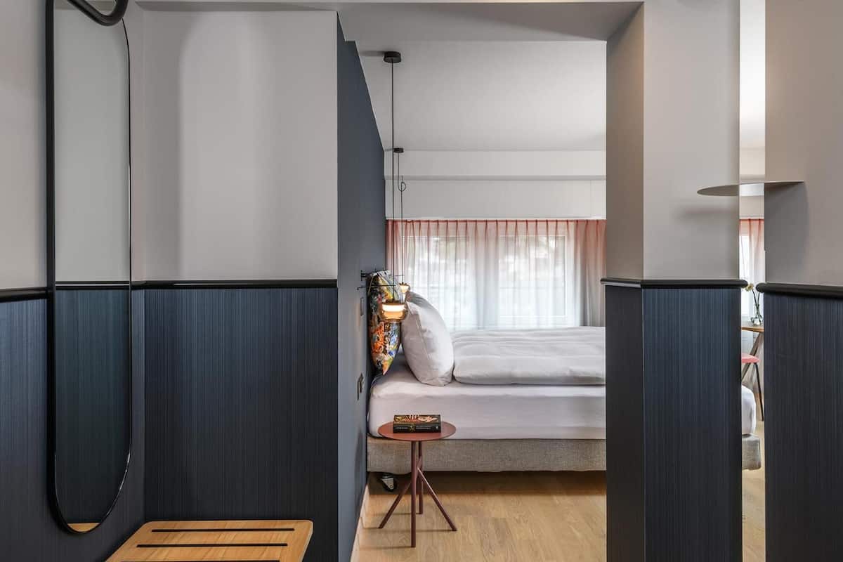 A modern hotel room with a bed, bedside table, and a small table with a lamp.