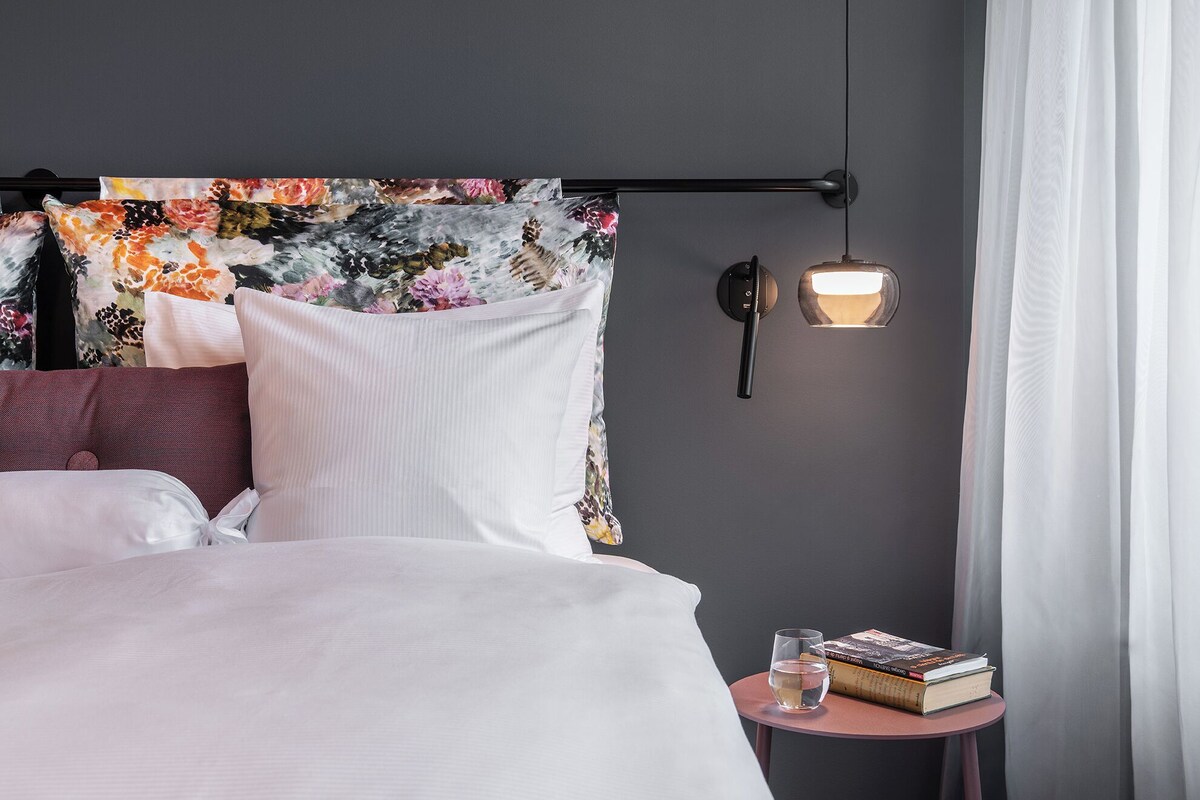 A neatly made bed with white linens, a floral headboard, a bedside table with a book and a glass of water, and a wall-mounted light fixture.