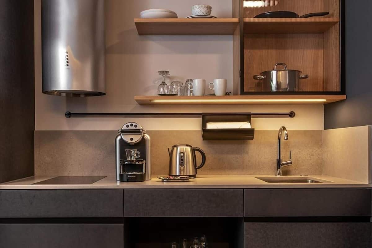A modern kitchen with a built-in stove, a coffee machine, and a sink. There are shelves above the counter holding dishes and glasses.