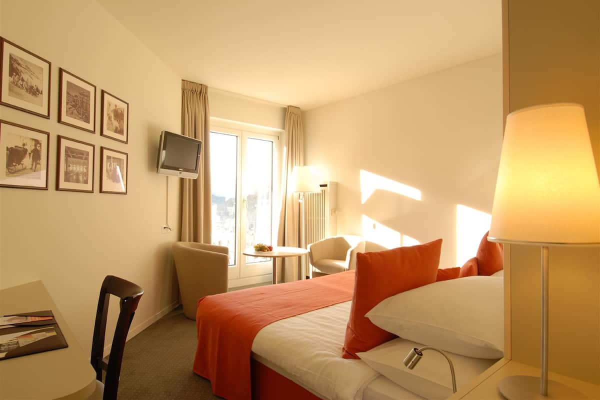 A hotel room with a bed, a desk, a chair, a television, and framed pictures on the wall.