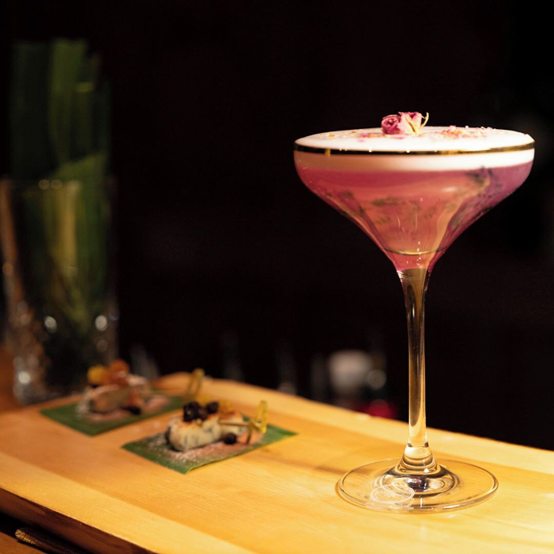 A martini glass with a pink hue on a wooden bar counter.