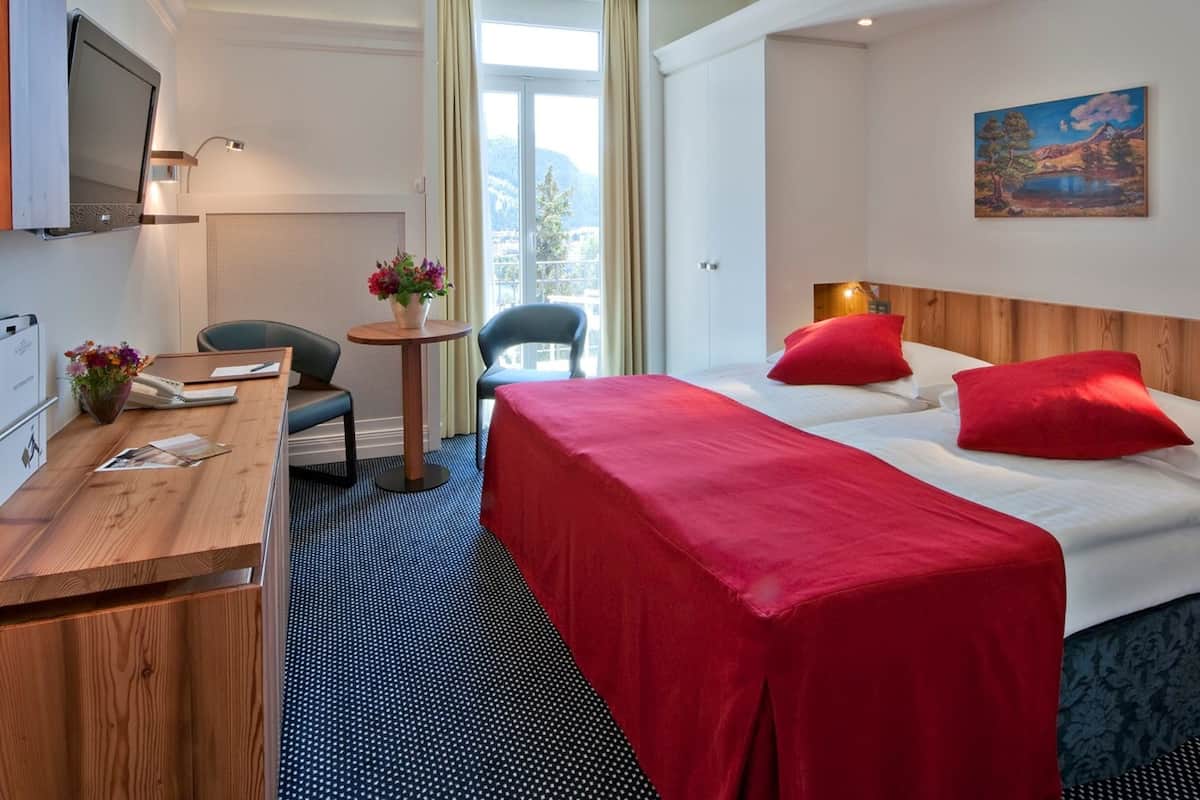 A hotel room with a bed, a desk, a chair, a small table with flowers, and a painting on the wall.