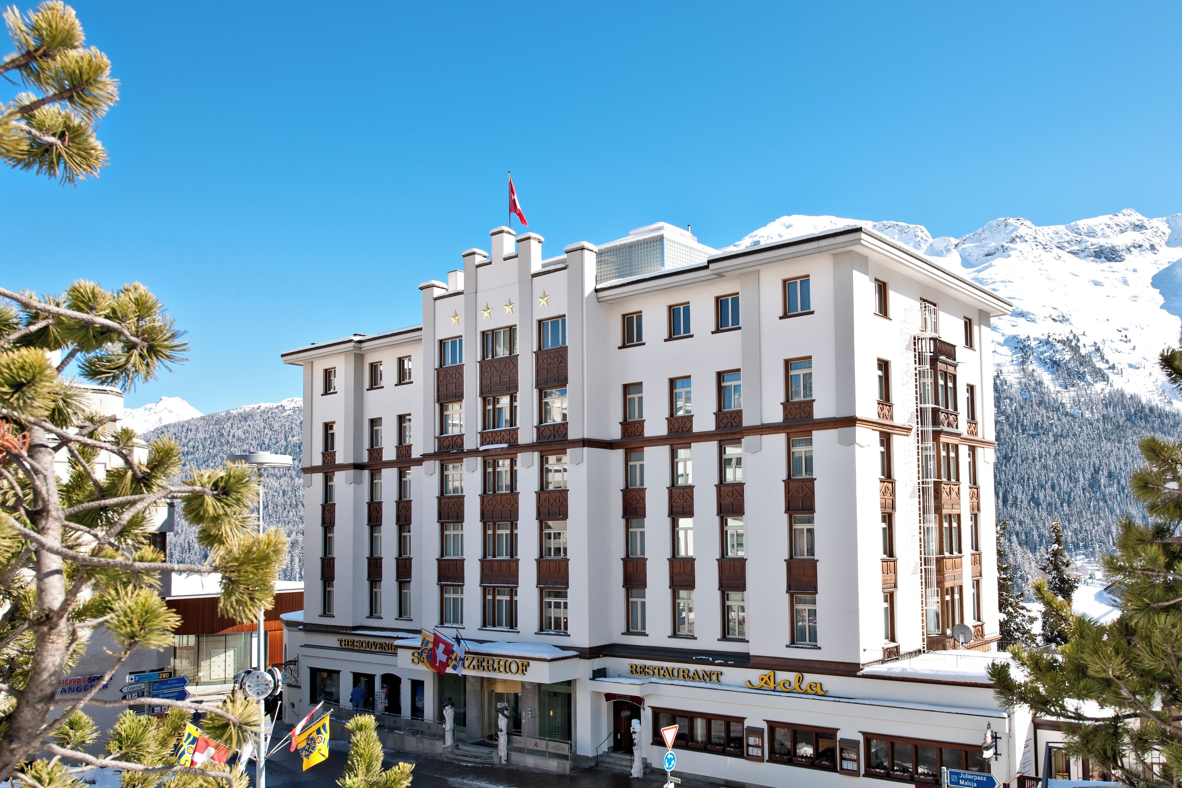 Un hotel de varios pisos con entrada central, banderas y montañas al fondo.