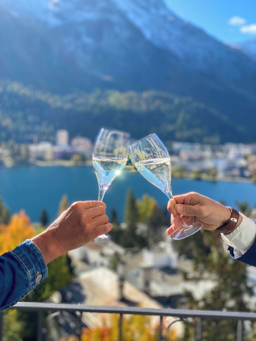 Dos personas brindan con copas de champán con un hermoso paisaje de un lago y montañas al fondo.