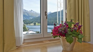 A room with a large window overlooking a lake and mountains. There is a potted plant with flowers on the windowsill. - Schweizerhof Swiss Quality Hotel (St. Moritz)