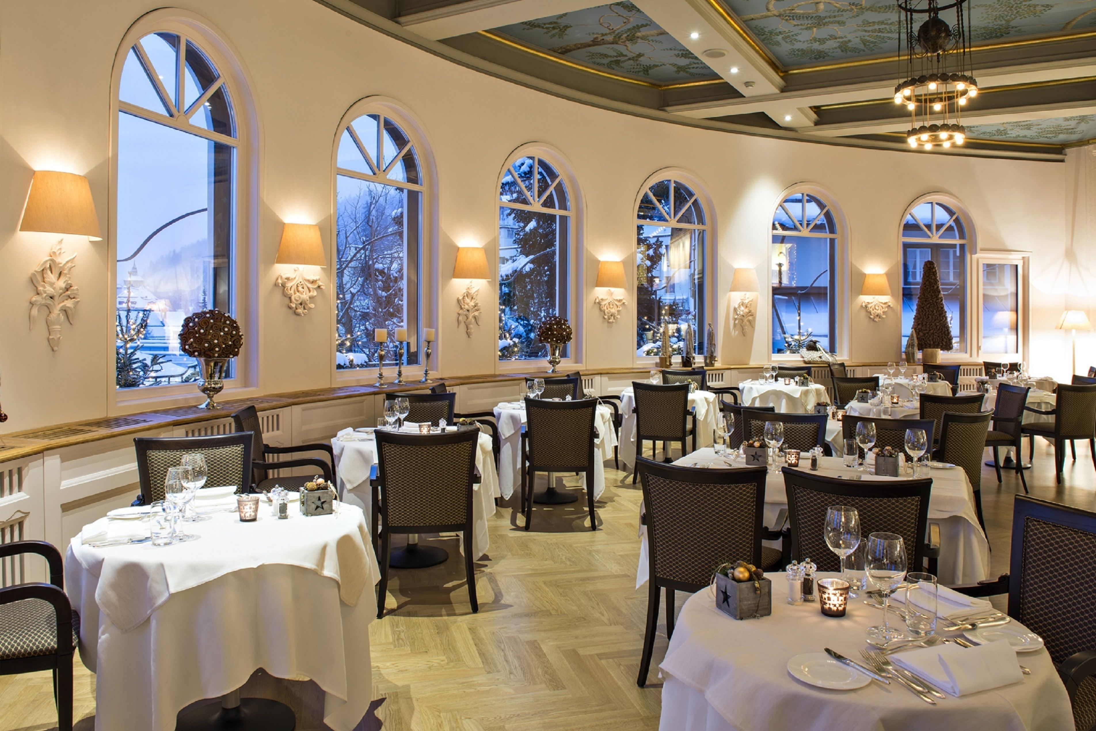 A restaurant with multiple tables set for dining, large arched windows, and chandeliers.