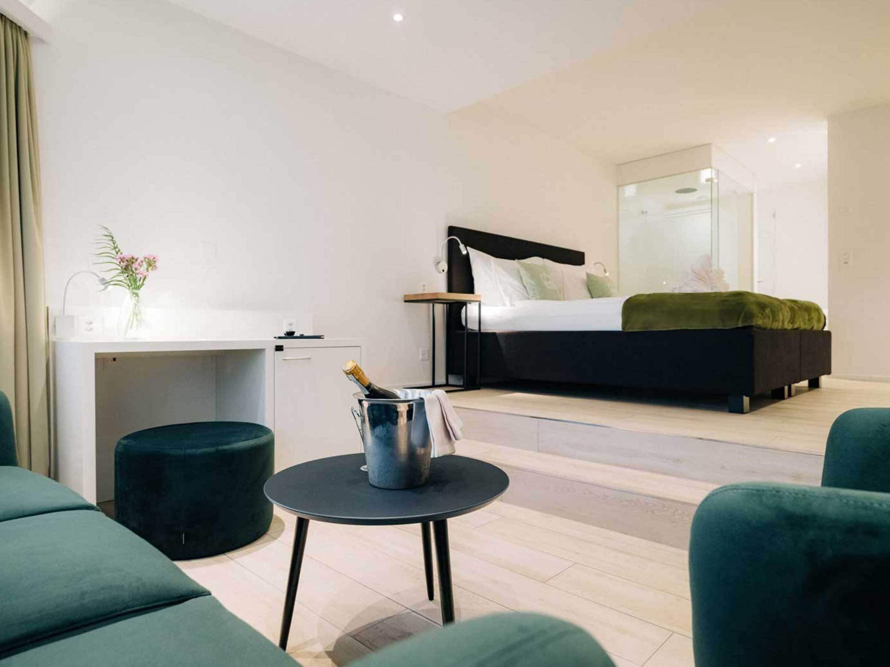A modern hotel room with a bed, a desk, a small table with a bottle, and green upholstered seating.