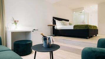 A modern hotel room with a bed, a desk, a small table with a bottle, and green upholstered seating.