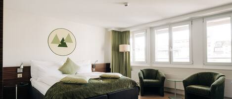 A modern hotel room with a bed, two armchairs, a small table, and a window with curtains.