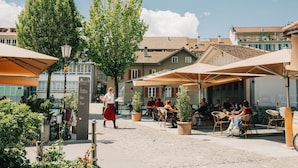 An outdoor café with tables and chairs under umbrellas, surrounded by buildings and trees.