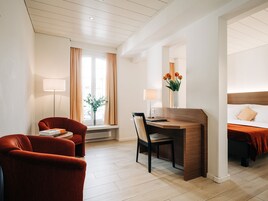 Una habitación de hotel con un escritorio de madera, un sillón rojo, una cama con cabecera de madera, una ventana con cortinas y un jarrón con flores.