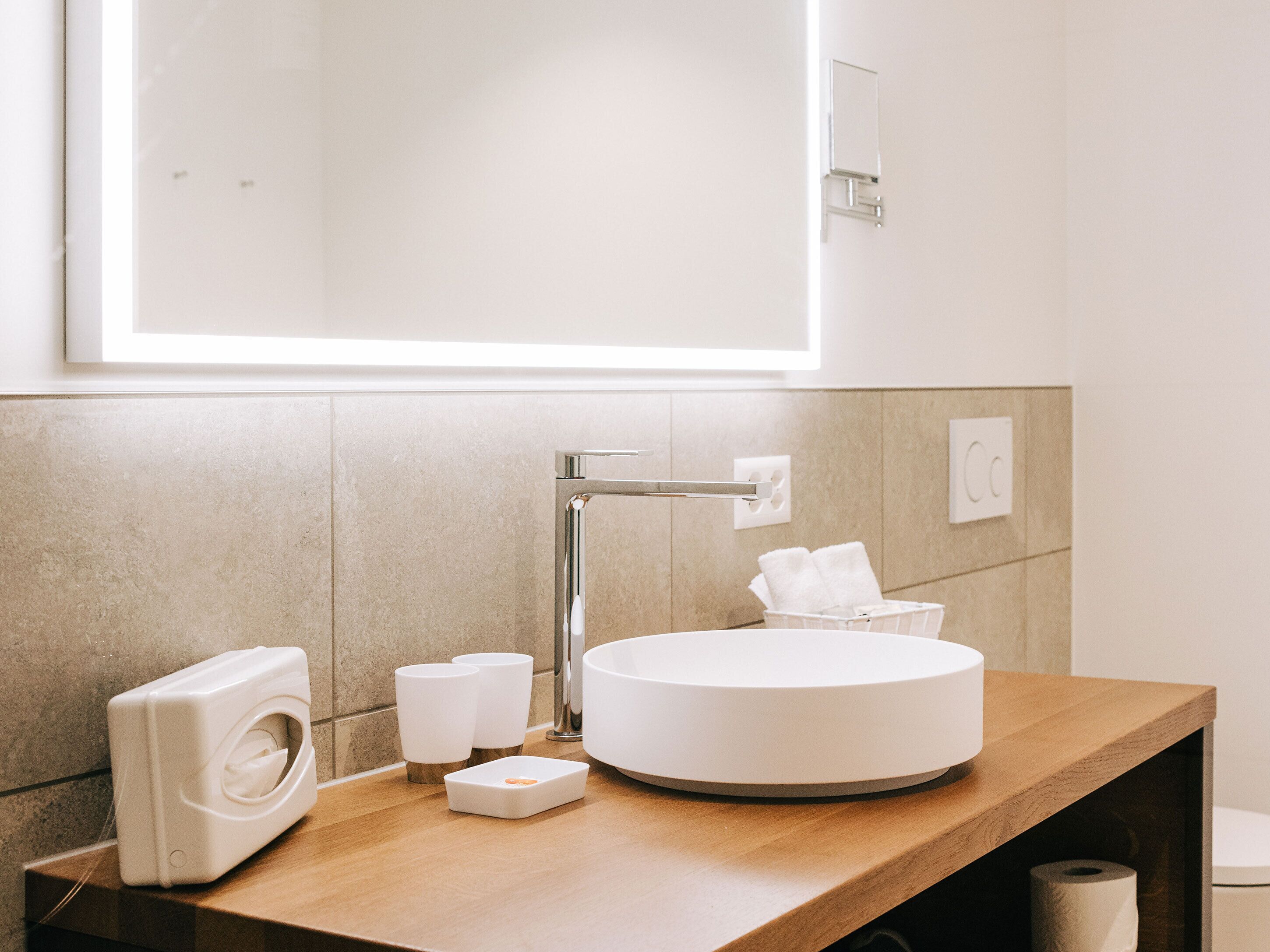 A modern bathroom with a round sink, a mirror, a towel dispenser, and a toilet paper holder.