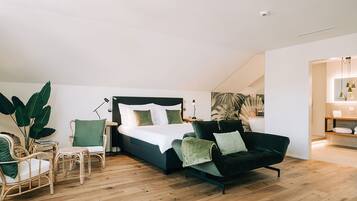 A modern bedroom with a bed, a sofa, and a chair. There is a large plant and a painting on the wall.