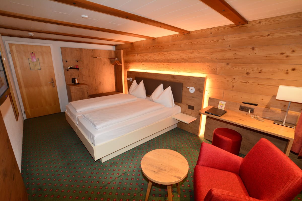 A hotel room with a large bed, a small table, a red chair, a desk with a lamp, and wooden walls.