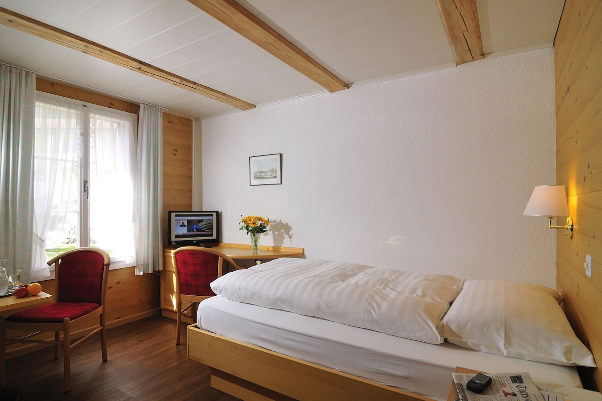 A hotel room with a wooden bed, a desk with a chair, a television, and a window with curtains.