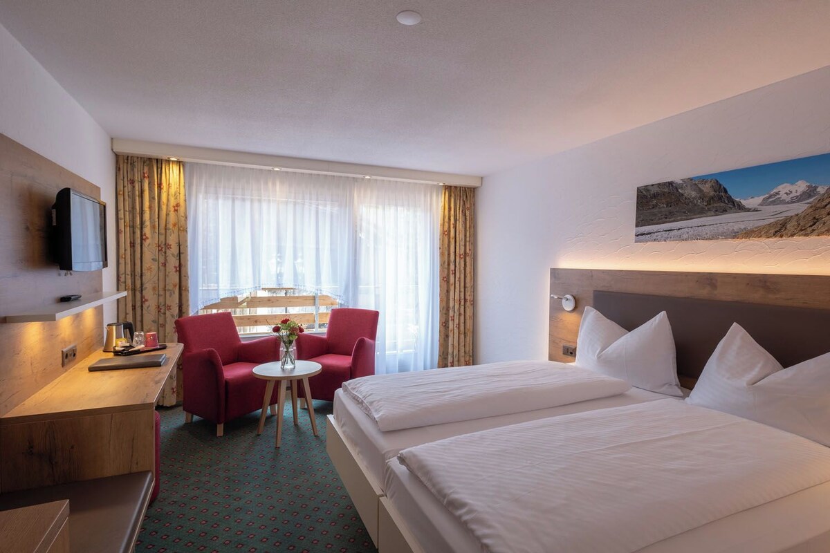 A hotel room with a bed, a desk with a chair, a small table with a vase, and a painting on the wall.