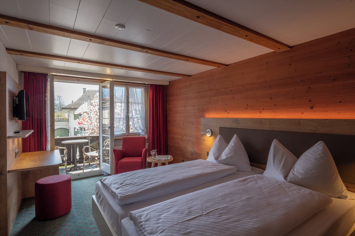 A hotel room with two beds, a wooden ceiling, a red chair, a small table, and a TV on a shelf.