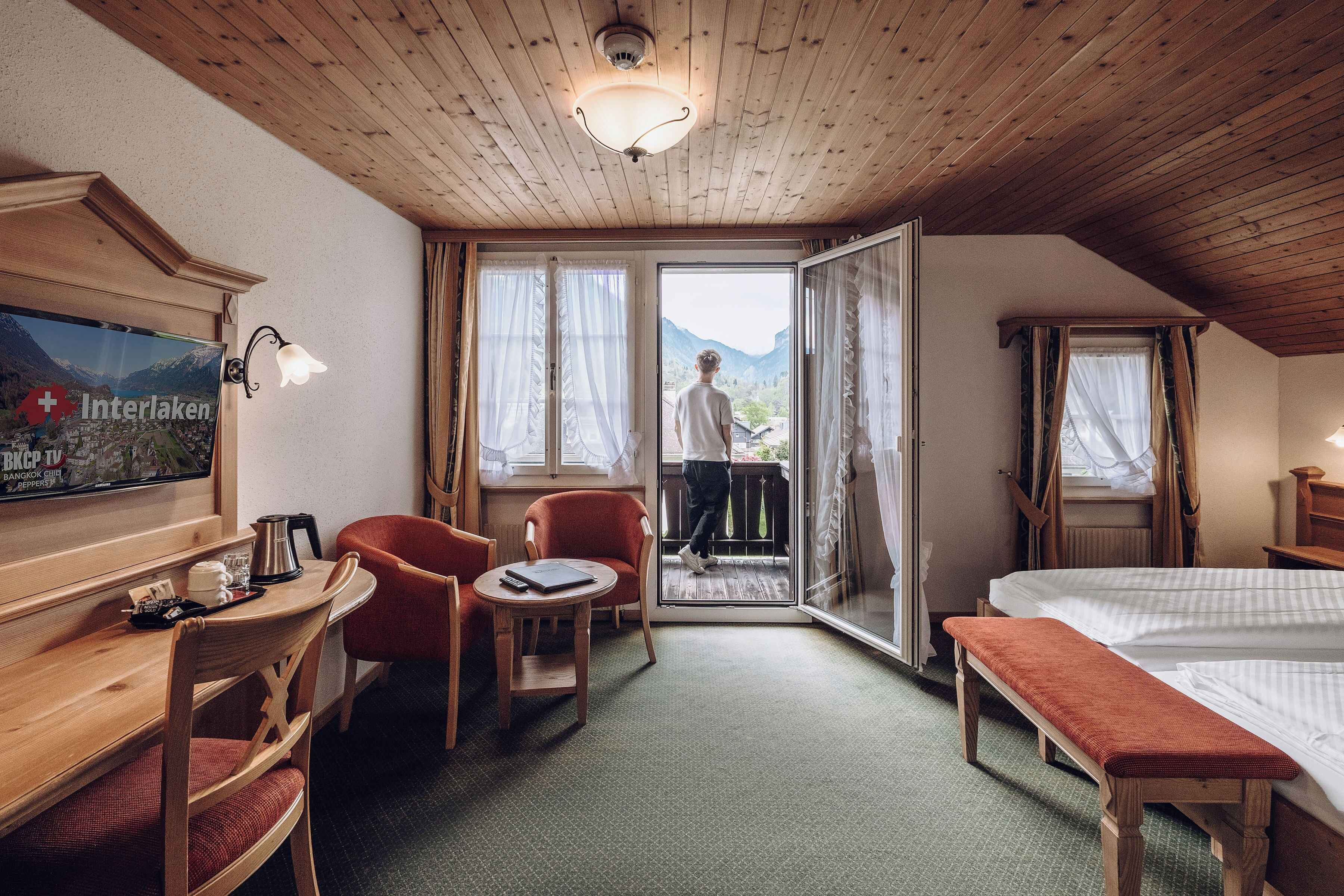 A hotel room with a wooden ceiling, a bed, a desk with a chair, a small table, and a view of mountains through the door.