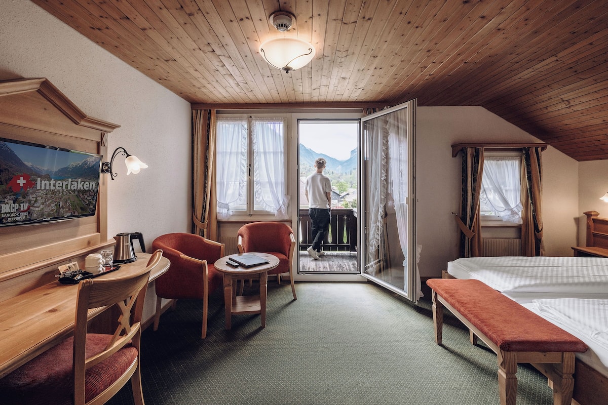 A hotel room with a wooden ceiling, a bed, a desk with a chair, a small table, and a view of mountains through the door.