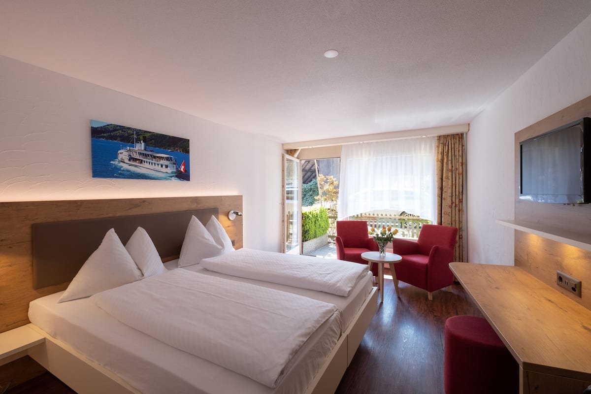 A hotel room with two beds, a red chair, a small table, a TV, and a painting of a boat on the wall.