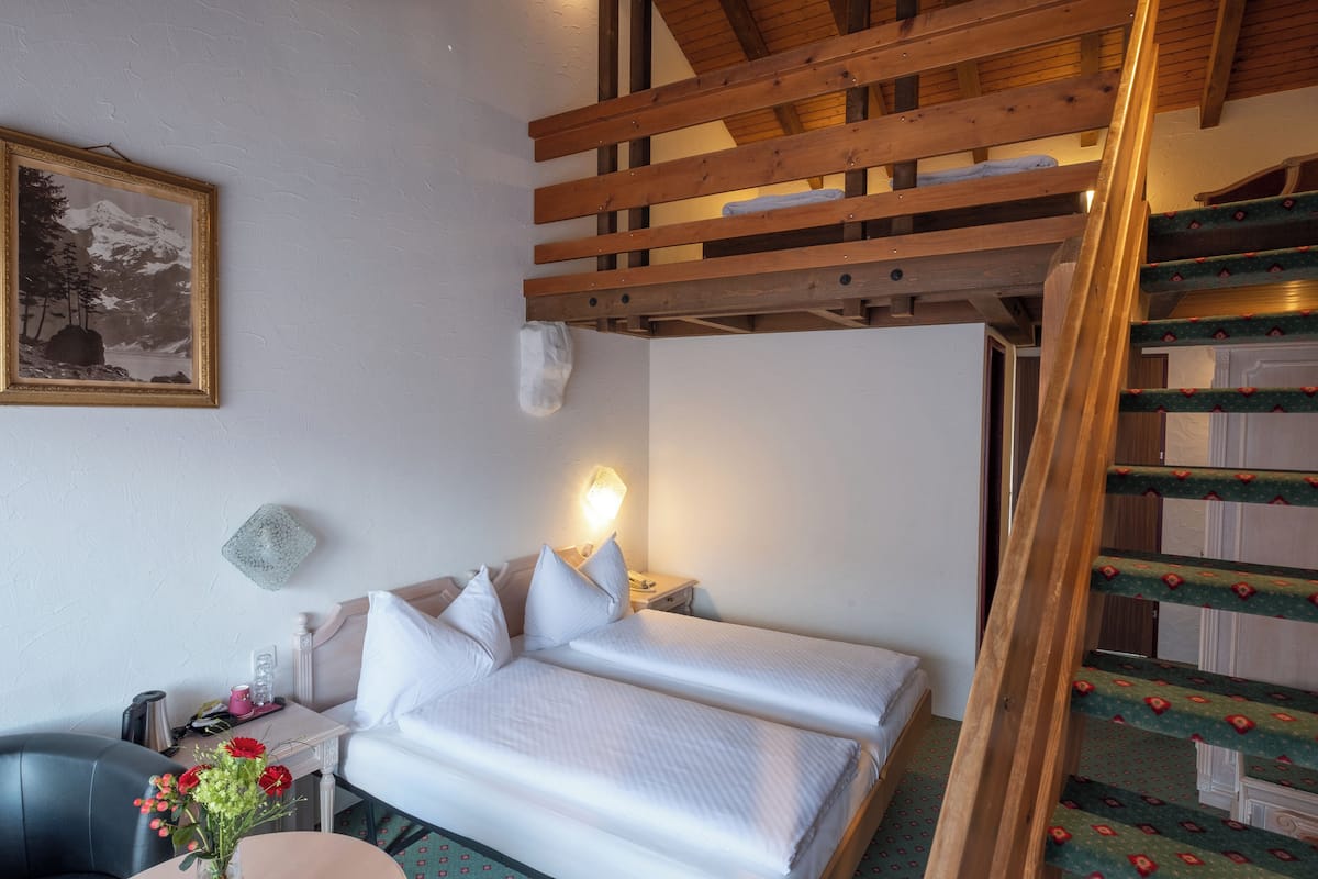 A hotel room with a bed, a nightstand, a chair, a desk, a vase with flowers, a framed picture on the wall, and a staircase.