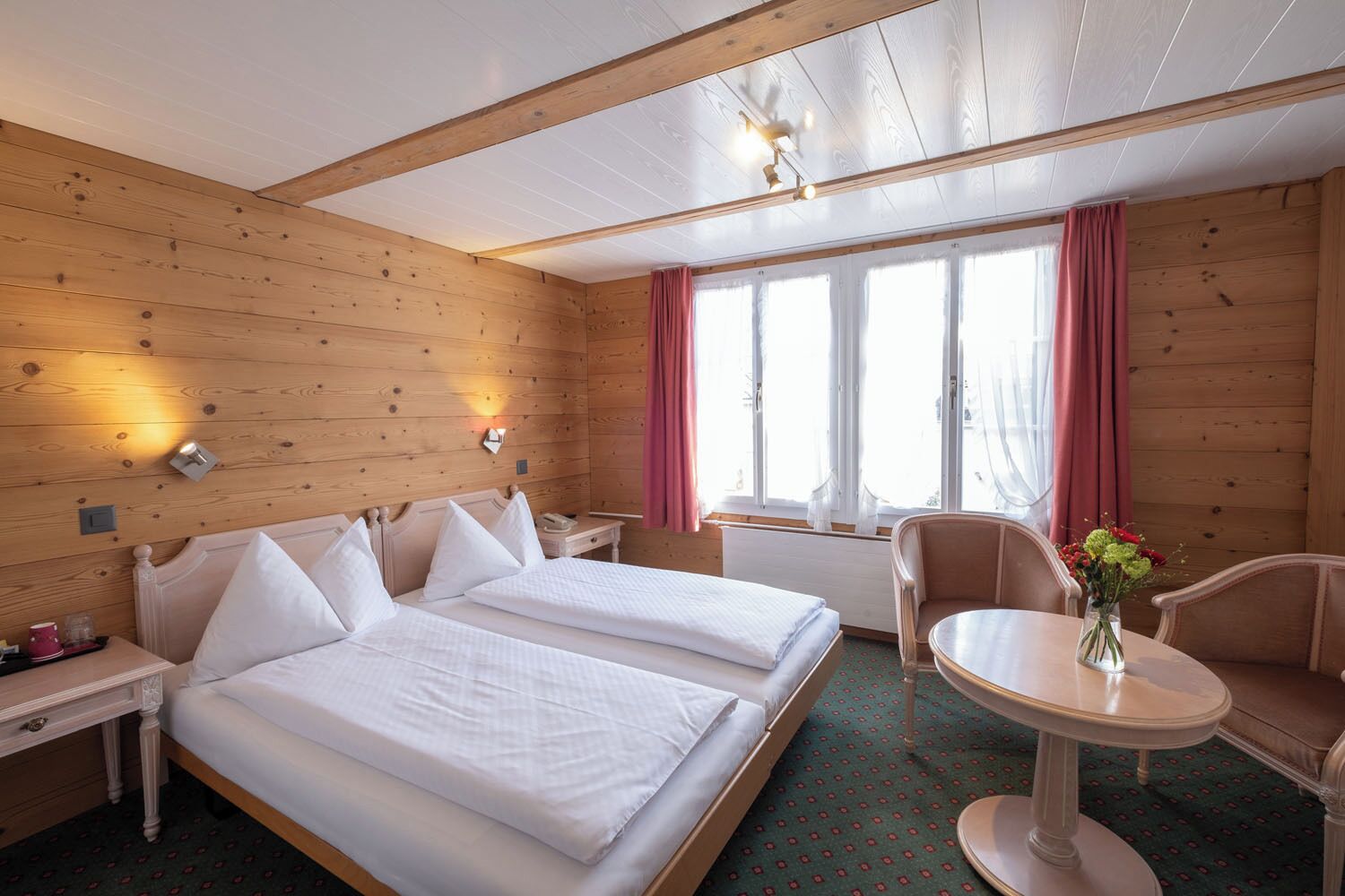 A hotel room with two beds, a small table, a chair, a window with curtains, and a wall-mounted light fixture.