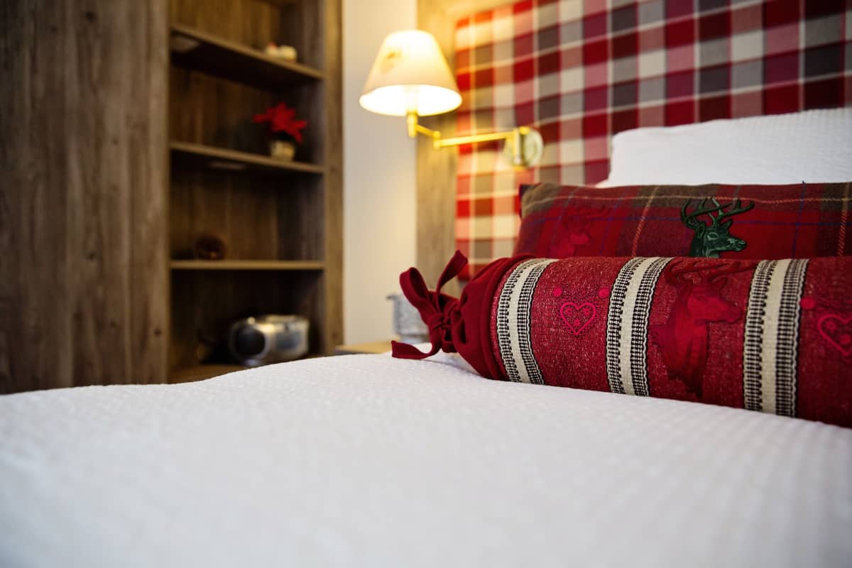A neatly made bed with a plaid headboard, a lamp, and a shelf with a red flower.