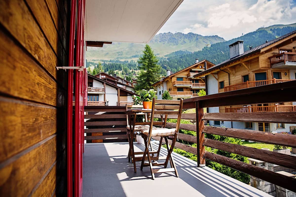 A wooden balcony with a table and chairs, overlooking a mountainous landscape with buildings in the background.