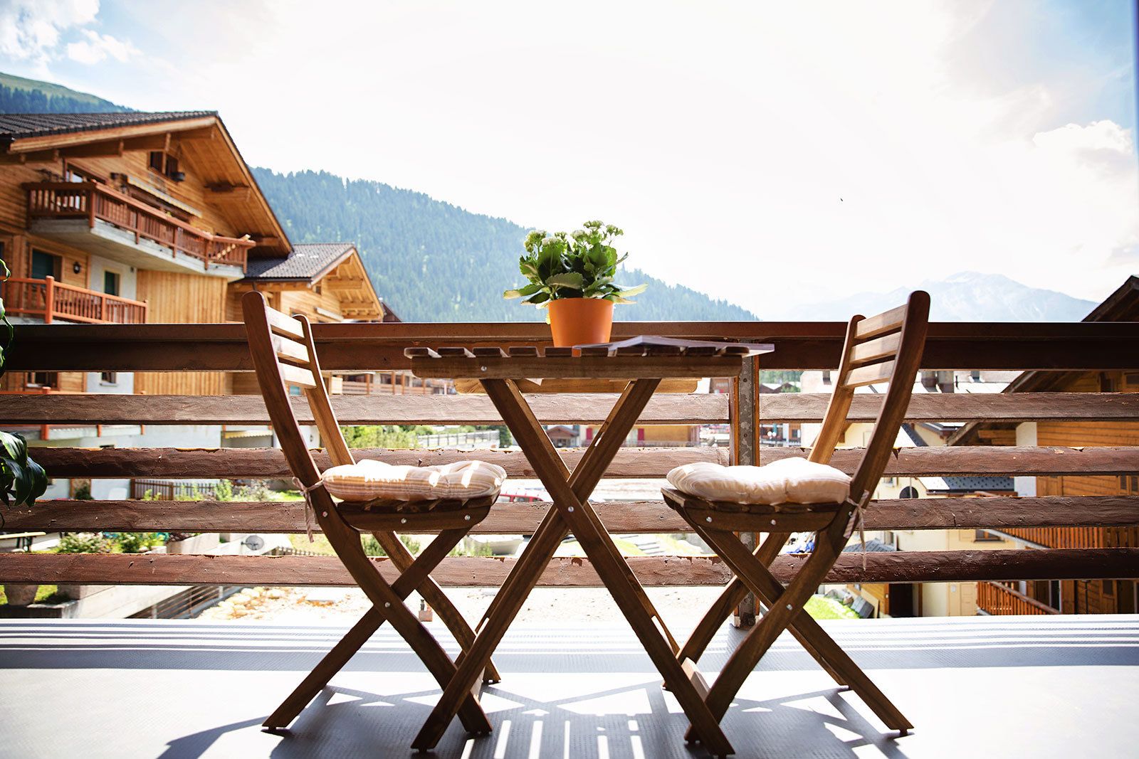 Una terraza de madera con una mesa, dos sillas, una planta en maceta y vistas a la montaña.