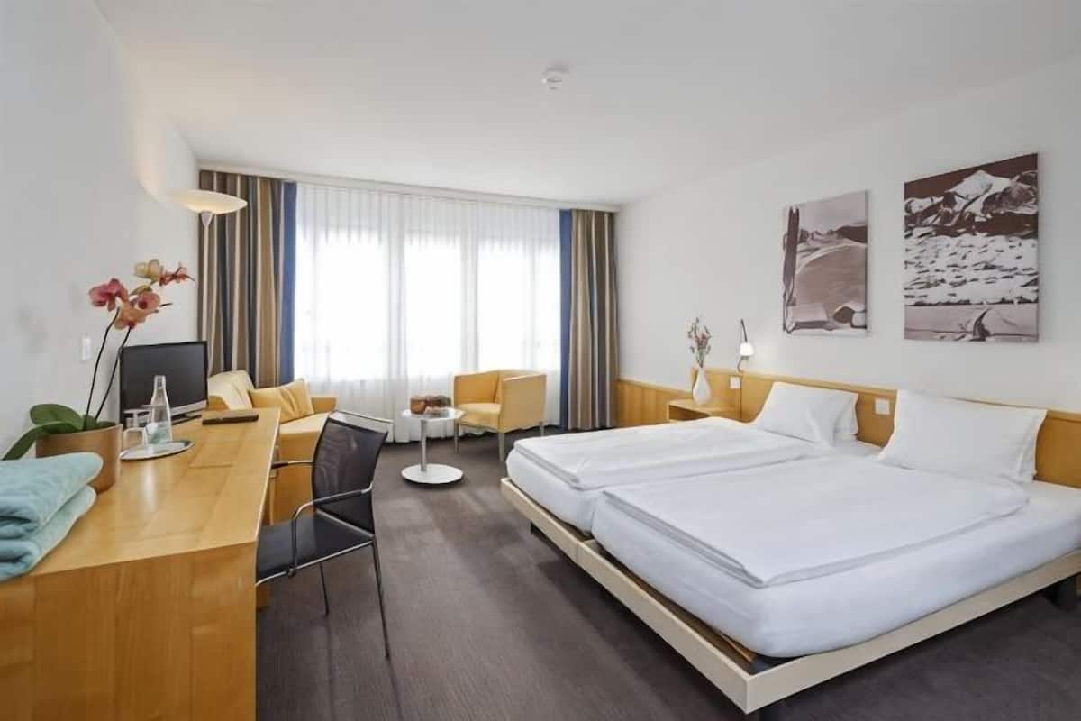 A hotel room with a bed, a desk, a chair, a small table, and a window with curtains.