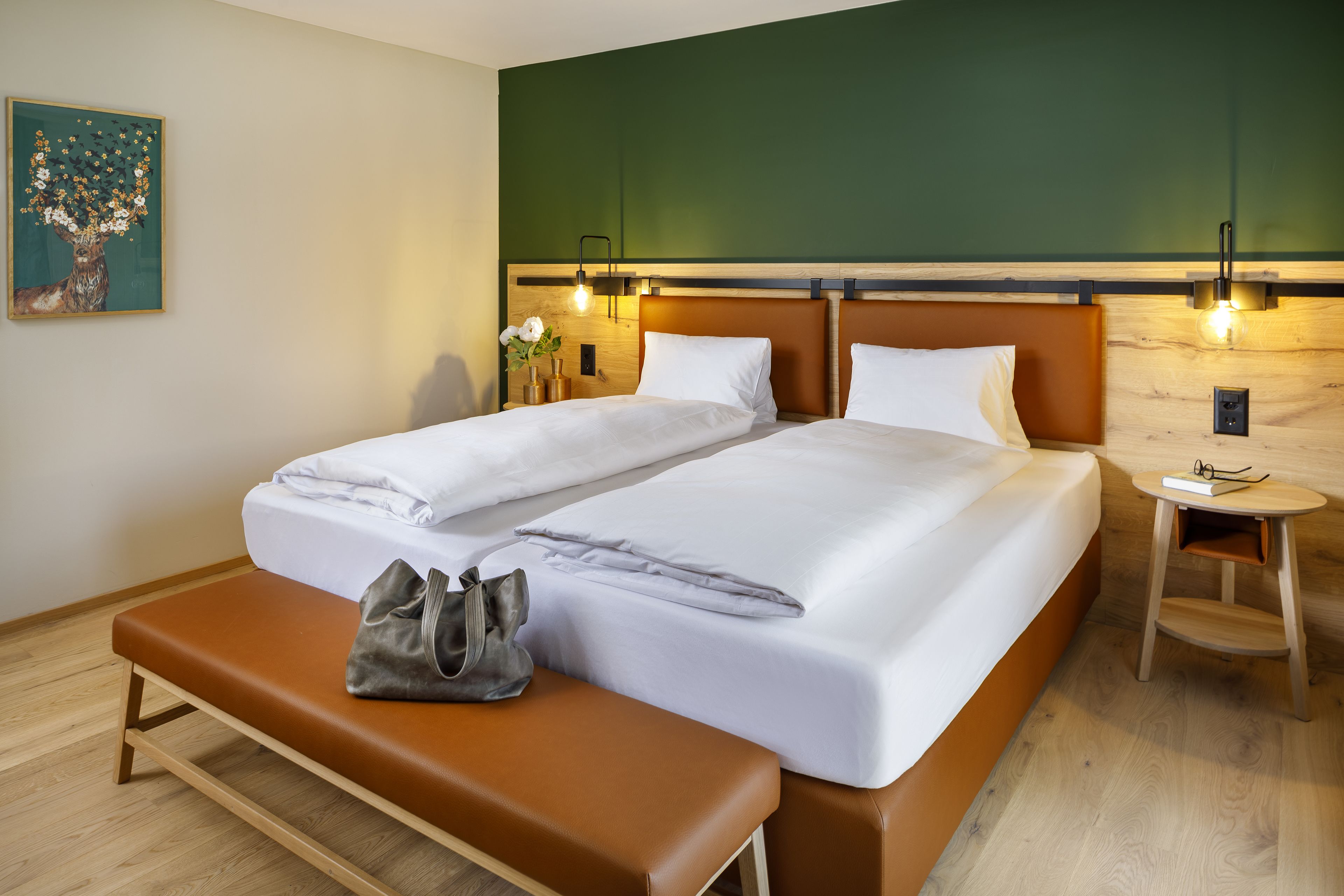 A double bed with white linens, a wooden bench, a bedside table with a lamp, and a painting on the wall.