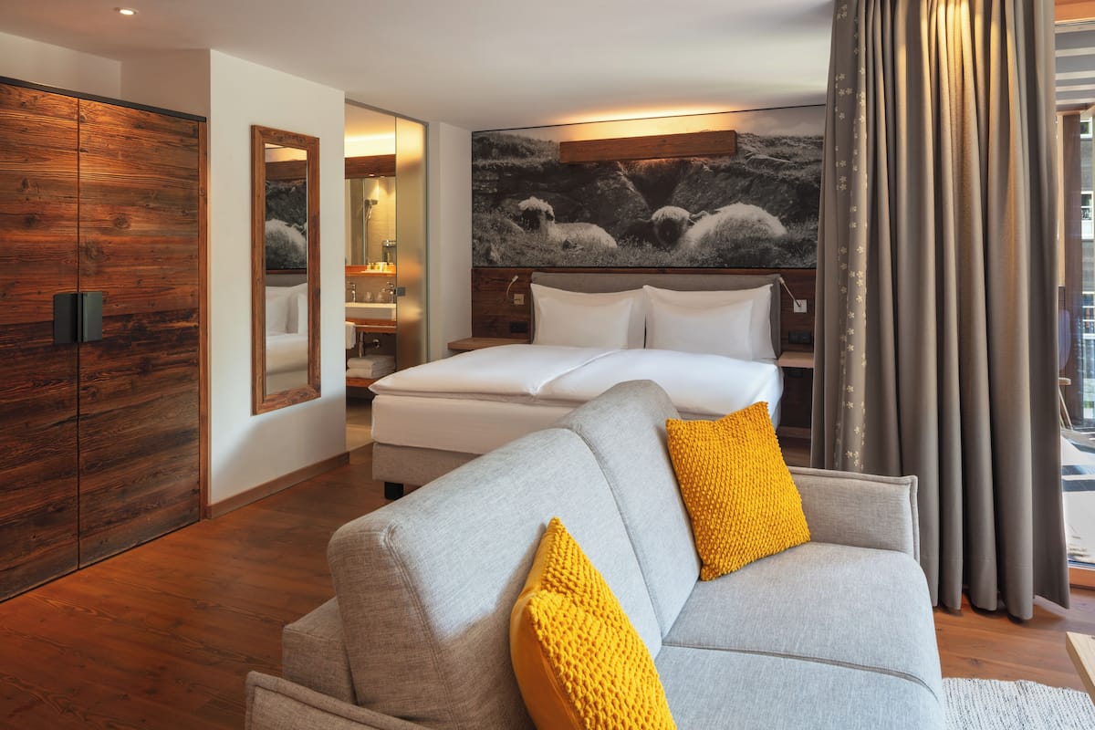 Habitació d'hotel moderna amb un sofà gris, un llit gran, un armari de fusta i una obra d'art penjada a la paret.