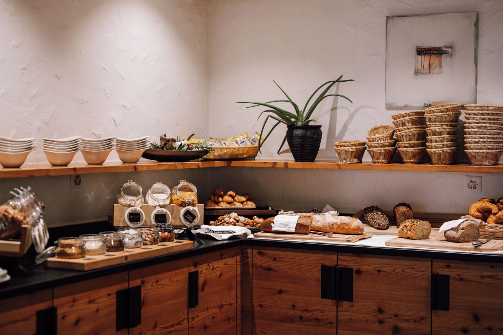 Un comptoir de boulangerie où l’on trouve une variété de pains et de viennoiseries, des pots de confitures et de pâtes à tartiner, ainsi que des paniers en osier.