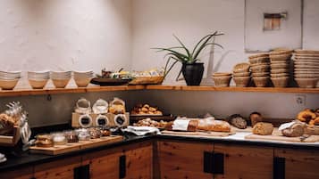 Un comptoir de boulangerie où l’on trouve une variété de pains et de viennoiseries, des pots de confitures et de pâtes à tartiner, ainsi que des paniers en osier.