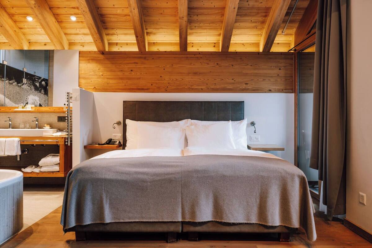 Habitació d'hotel amb sostre de fusta, un llit gran amb capçaleri fosc, una tauleta de nit amb una làmpada, un bany amb un lavabo i mirall, i una finestra gran amb cortines.