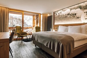 Habitación de hotel con una cama grande, un escritorio con silla, una mesita y vistas a las montañas desde la ventana.
