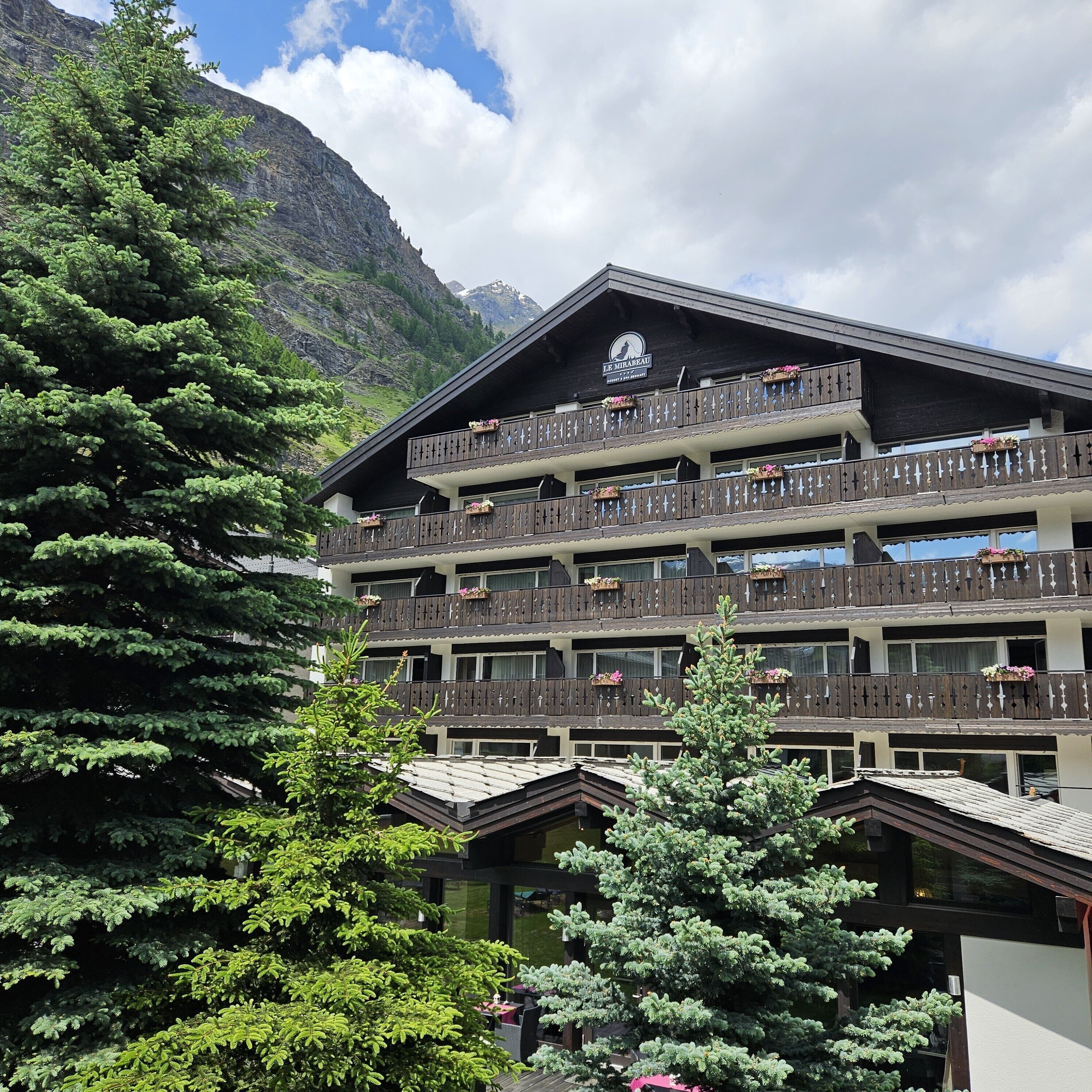 A multi-story building with balconies, surrounded by trees and mountains.