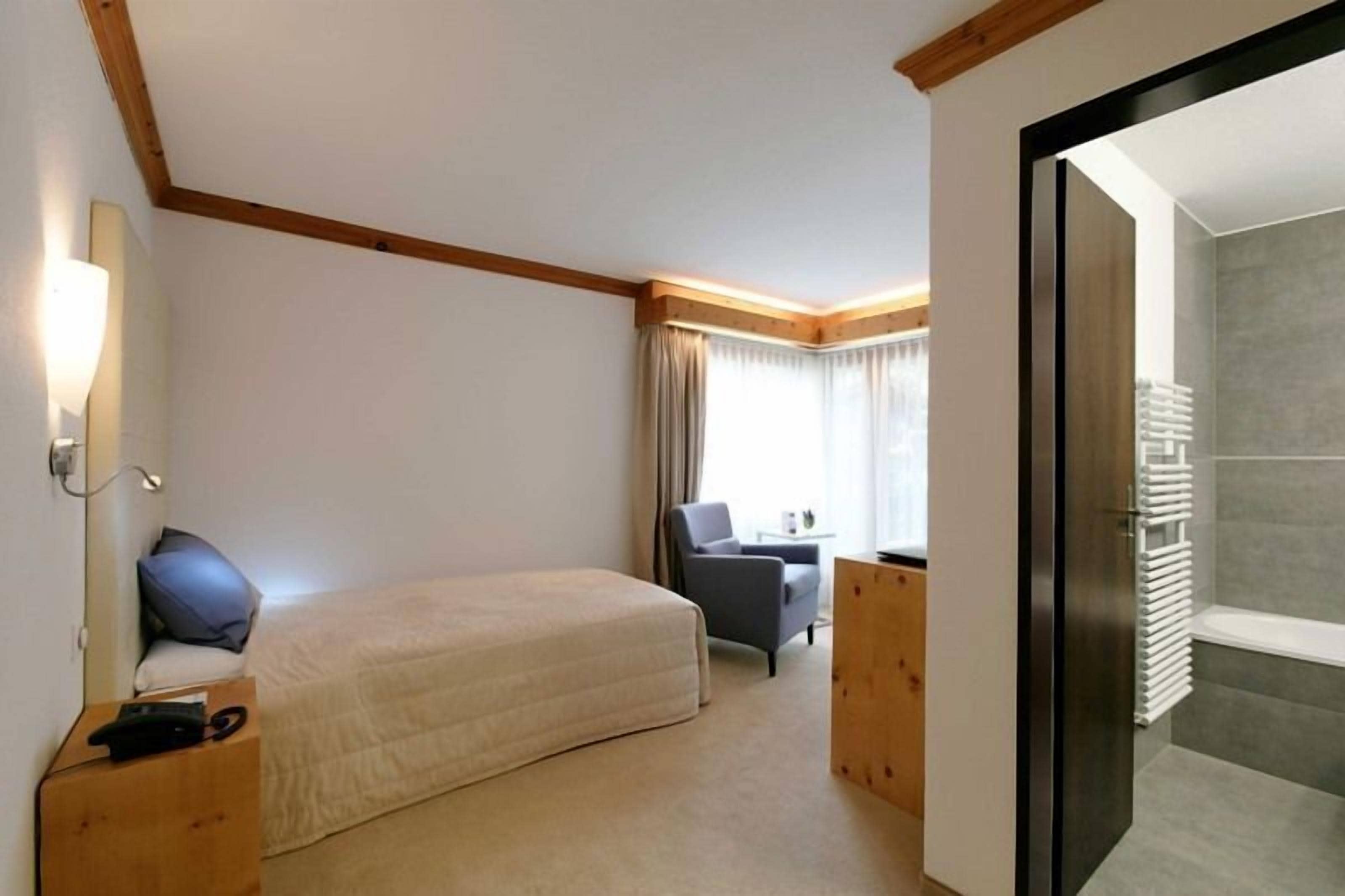A hotel room with a bed, a desk, a chair, a window with curtains, and a bathroom with a towel rack.