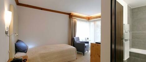 A hotel room with a bed, a desk, a chair, a window with curtains, and a bathroom with a towel rack.