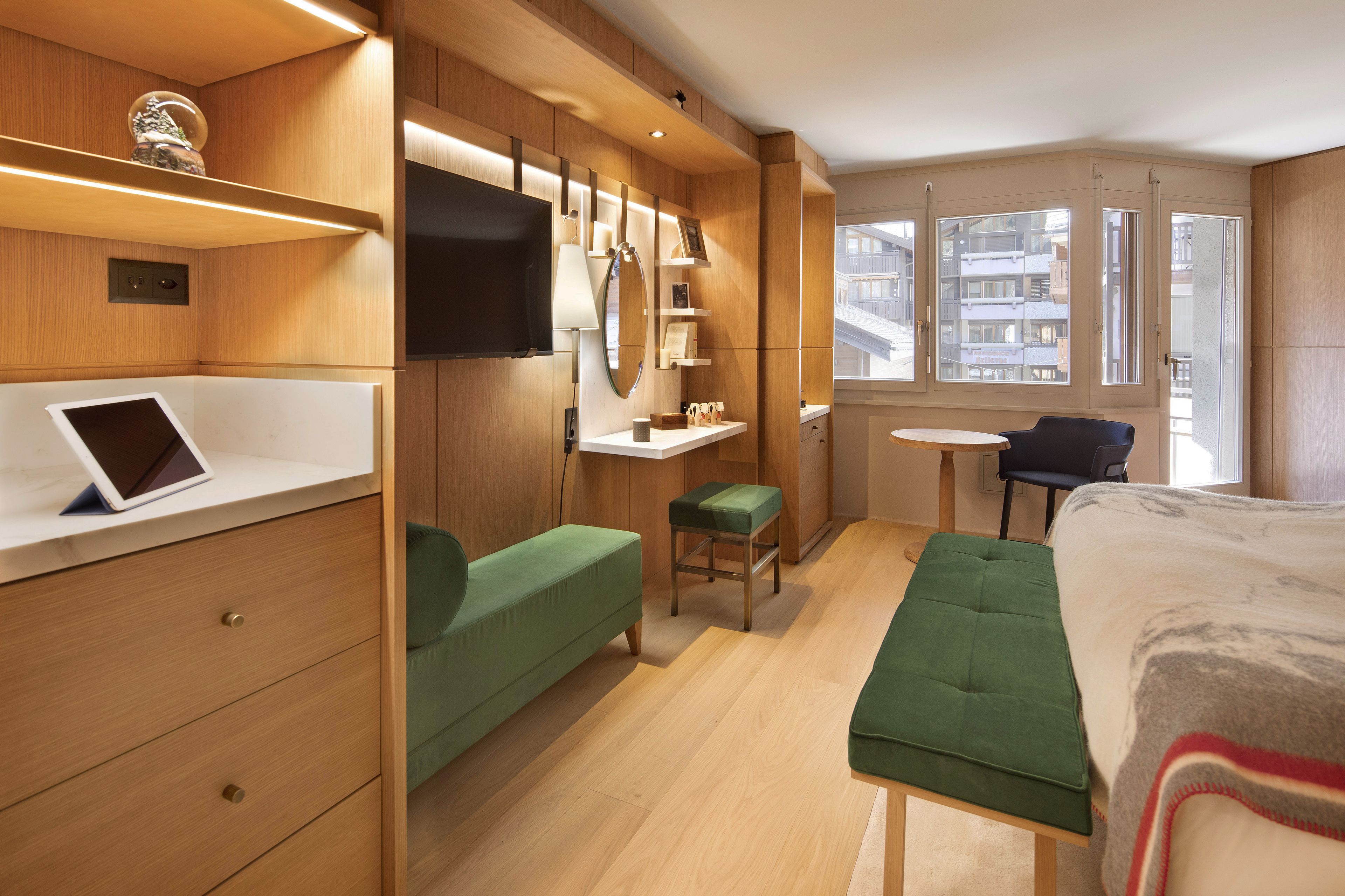 A modern hotel room with a bed, a desk, a chair, a green sofa, a small table, and a TV.