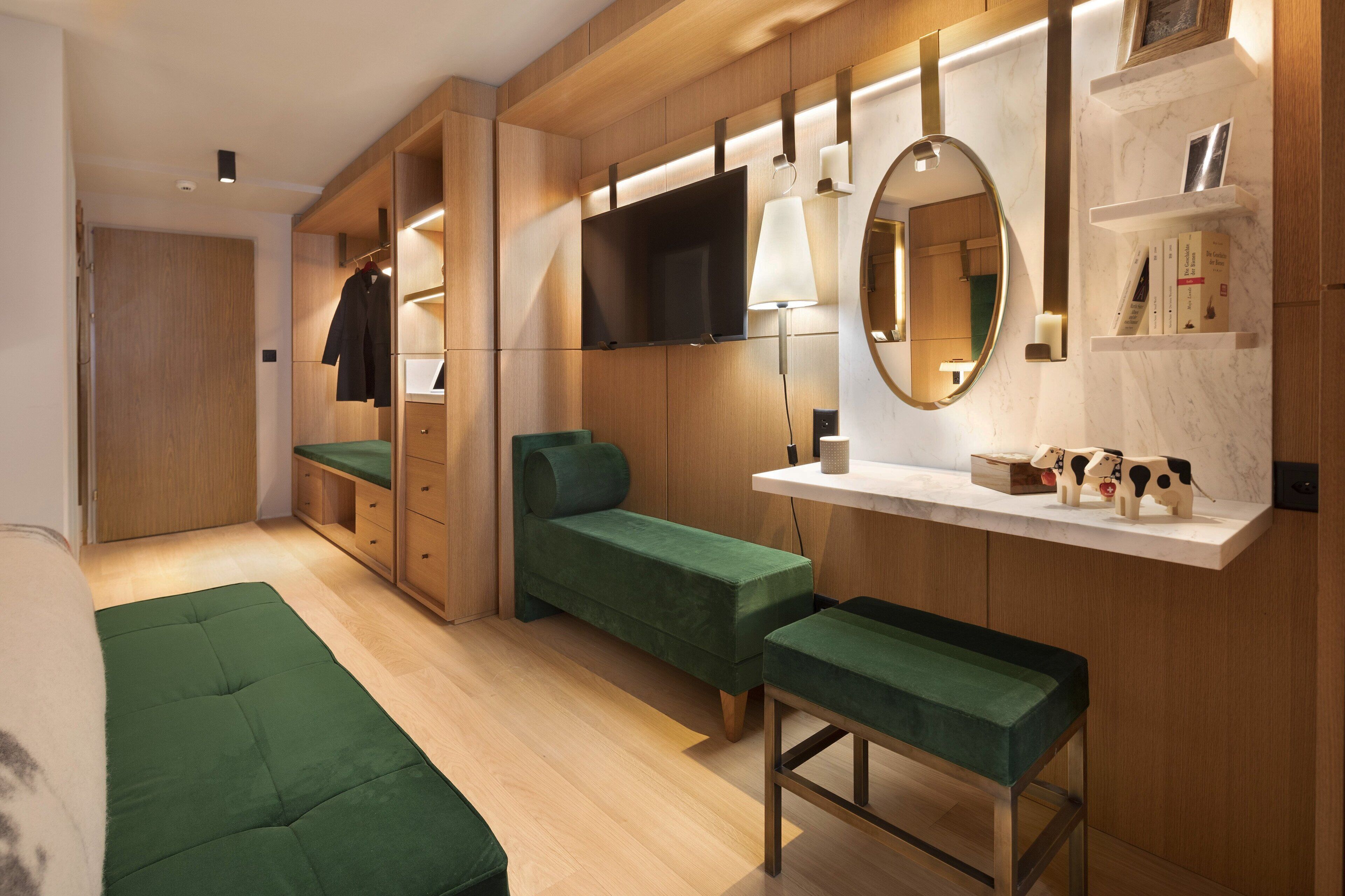A modern bedroom with a green bed, a bench, a TV, a mirror, and wooden shelving with books and a lamp.
