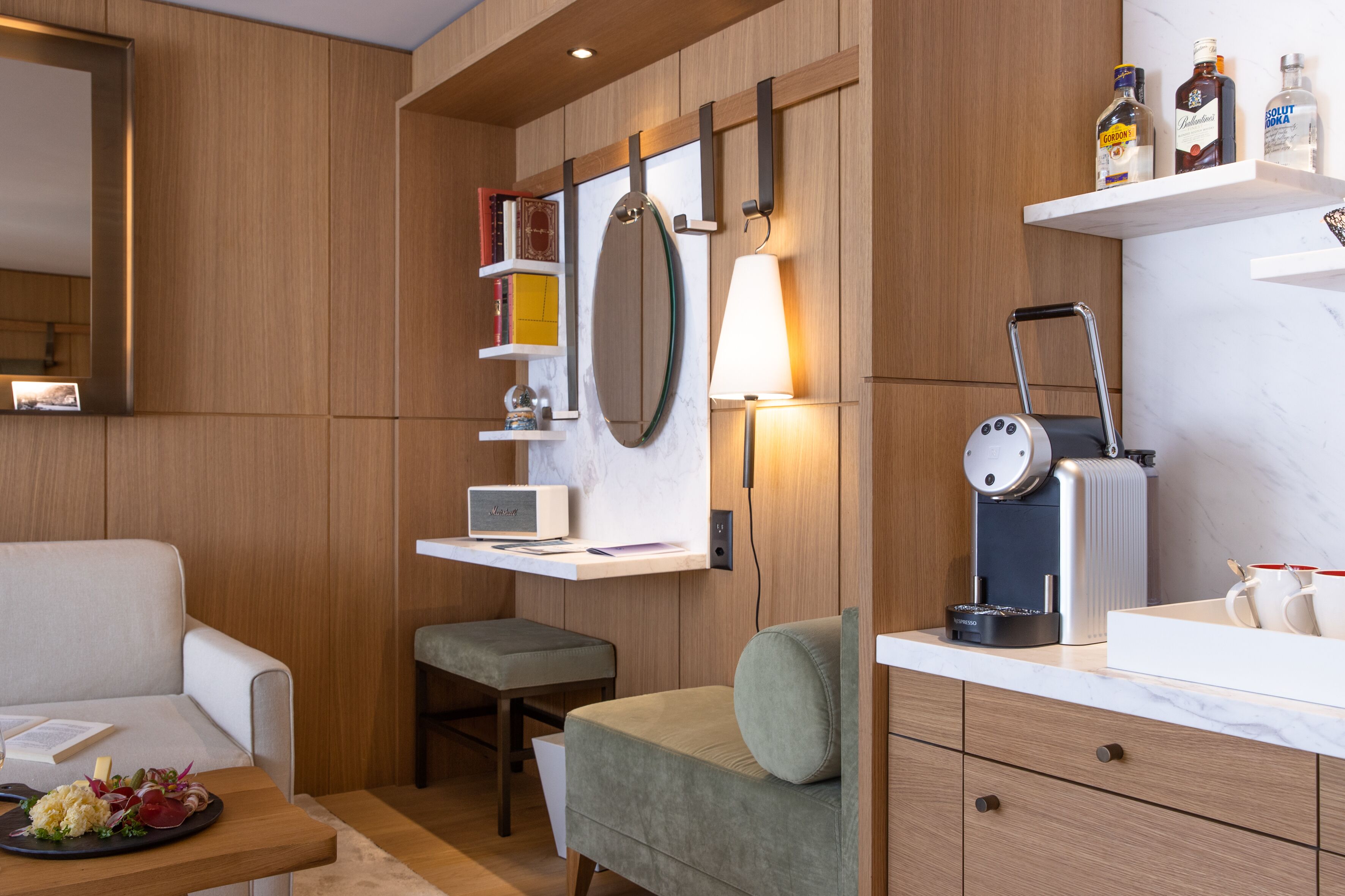 Une chambre d’hôtel moderne avec un mur lambrissé en bois, un canapé, une petite table sur laquelle se trouve une assiette de nourriture, une machine à café et une étagère avec des bouteilles.