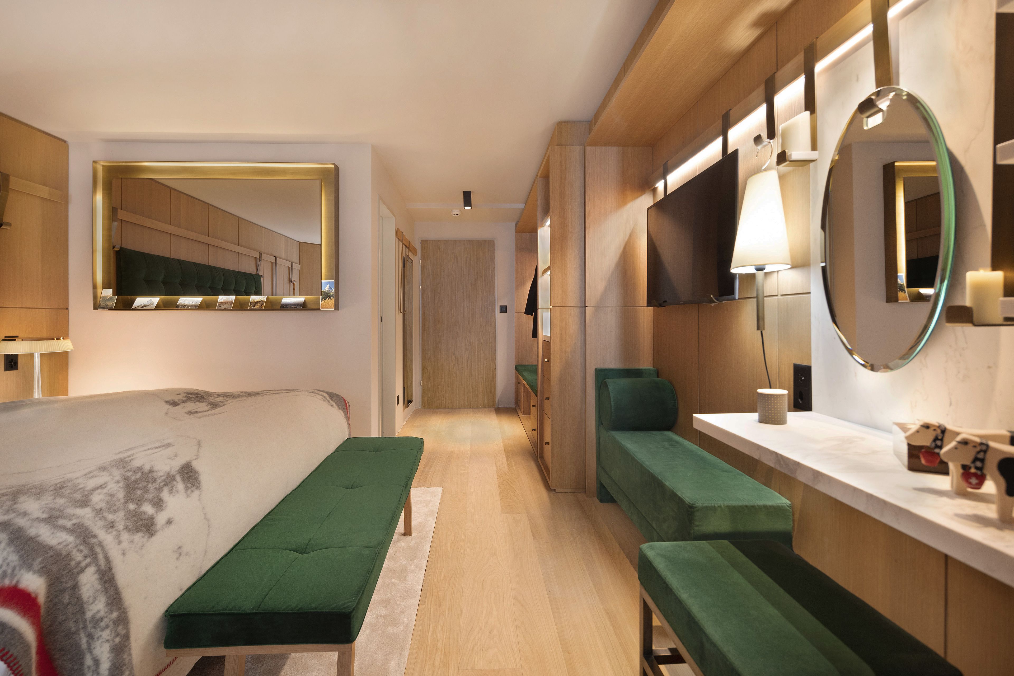 A modern hotel room with a large bed, green upholstered seating, a vanity with mirrors, and a wooden floor.