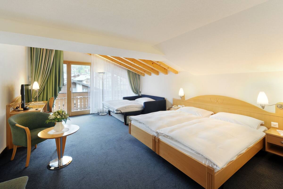 A hotel room with a large bed, a desk with a chair, a small table with a vase, and a view of the outside through a window with curtains.