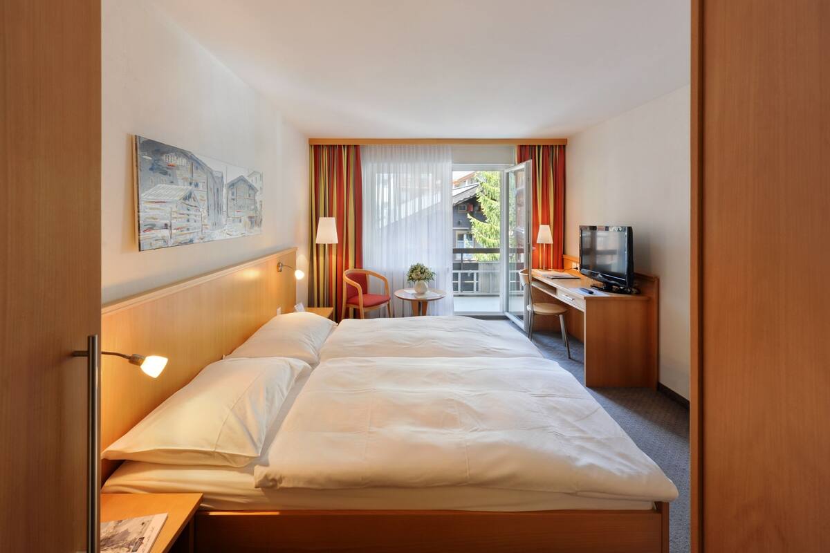 A hotel room with a large bed, a desk with a computer, a chair, a TV, and a window with curtains.