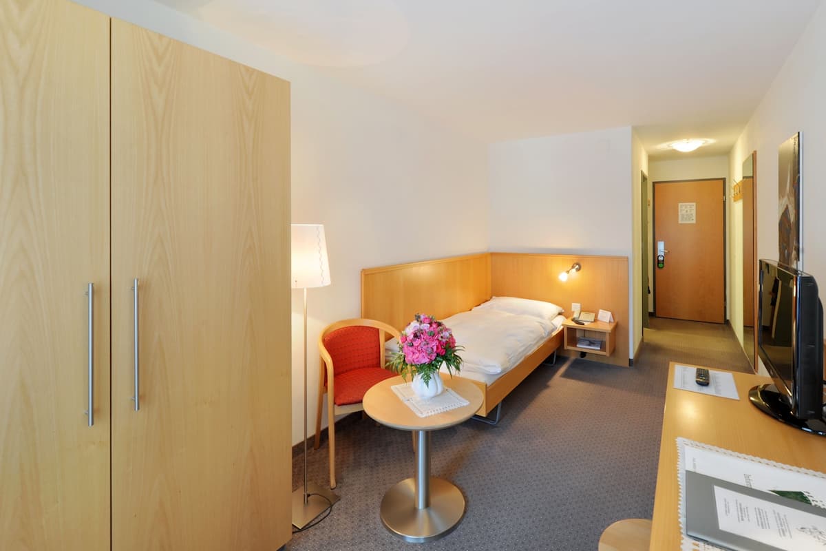 A hotel room with a bed, a desk with a lamp, a chair, a TV, and a wardrobe.