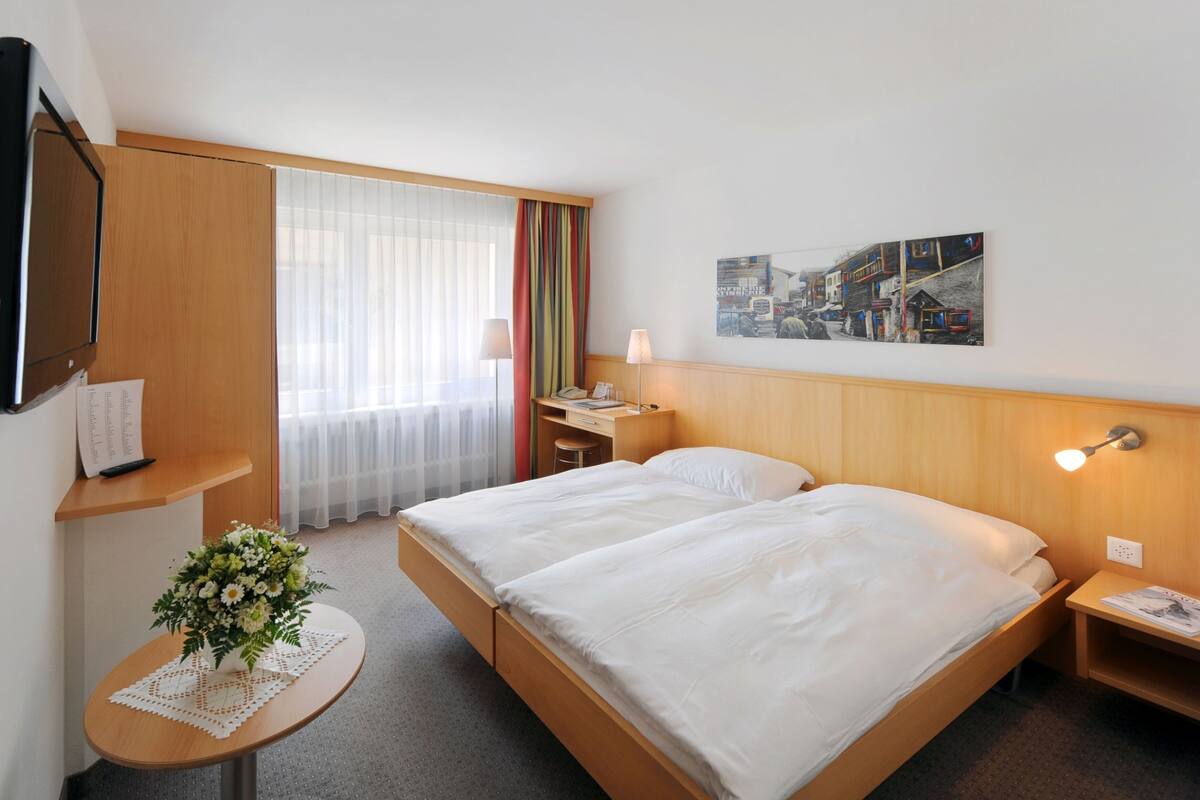 A hotel room with a double bed, a desk with a chair, a small table with a vase of flowers, a lamp, and a painting on the wall.