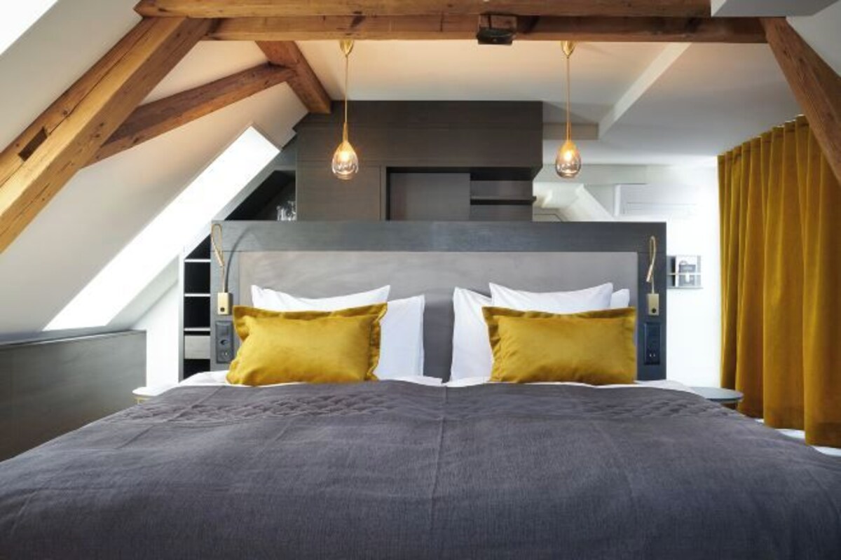 A bedroom with a sloped ceiling, a large bed with gray bedding, two mustard pillows, and pendant lights.